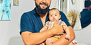 Thabit is registered to the contest to win money with this photo: man, baby, indoor, smile, beard, holding, mirror, painting, decor, casual_clothing, child, portrait, happy, furniture, book, bracelet, shorts, striped_shirt, reflection, family