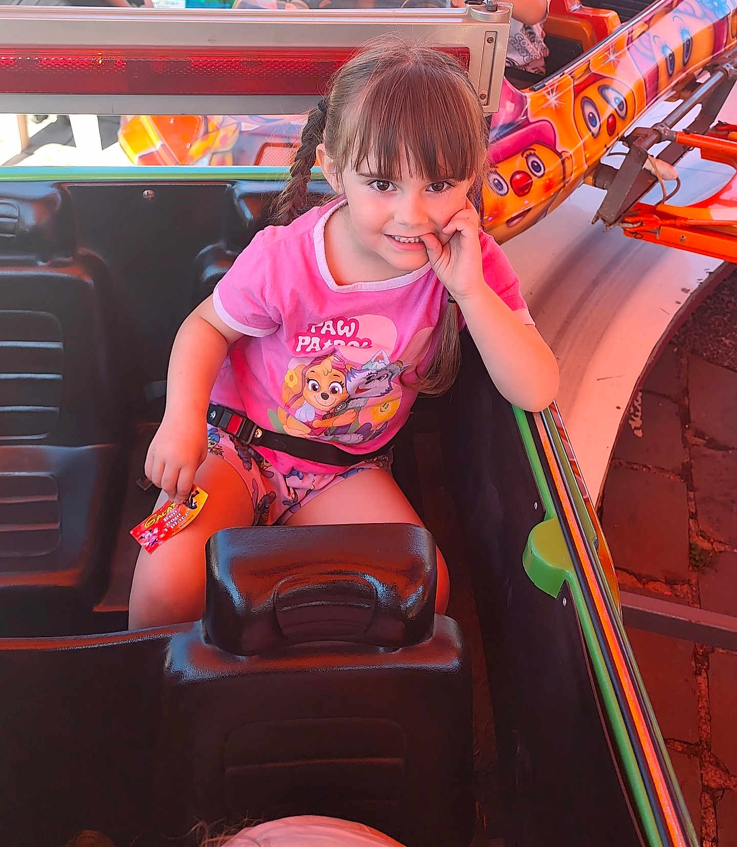 Capucine participe au concours pour gagner de l'argent avec cette photo : child, girl, braids, pink_clothing, paw_patrol, amusement_ride, seatbelt, ride_car, colorful, playful, smiling, finger_in_mouth, card, daylight, outdoor, entertainment, fun, seat, person, hair