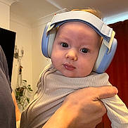 August is registered to the contest to win money with this photo: adult_hand, baby, child, clothing, curious, curtain, ears, expression, face, head, headphones, holding, indoor, light, long_sleeve, person, plant, portrait, room, wall