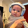 August is registered to the contest to win money with this photo: baby, child, headphones, person, indoor, holding, adult_hand, curious, face, clothing, long_sleeve, ears, head, room, curtain, plant, wall, light, portrait, expression