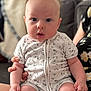 August is registered to the contest to win money with this photo: baby, infant, child, onesie, clothing, sitting, hand, face, expression, indoor, couch, blanket, person, skin, cheeks, curly_hair, holding, soft_lighting, floral_pattern, background