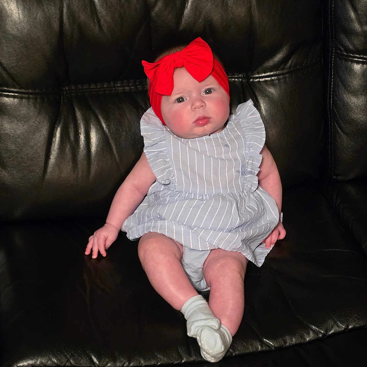Delylah is registered to the contest to win money with this photo: baby, black_leather_couch, casual, child, comfort, cute, expression, face, furniture, headband, indoor, infant, person, portrait, red_bow, sitting, skin, sock, striped_dress, young_child