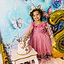 child, girl, birthday, cake, balloon, number_two, pink_dress, flower_crown, butterflies, decorations, smiling, happy, party, table, floral, background, indoor, curly_hair, footwear, celebration