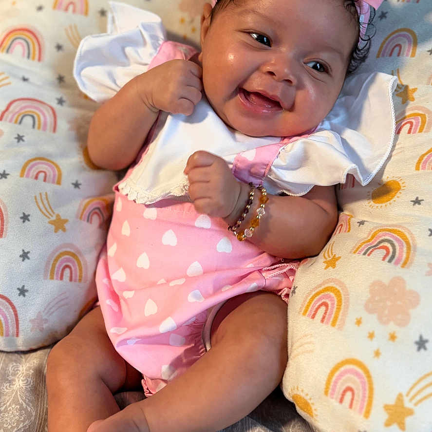 Azuri Howard is registered to the contest to win money with this photo: baby, bow, bracelet, child, close_up, cushion, cute, happy, headband, indoors, infant, lying_down, person, pink_clothing, rainbow_pattern, skin, smiling, soft_lighting, stars, toddler