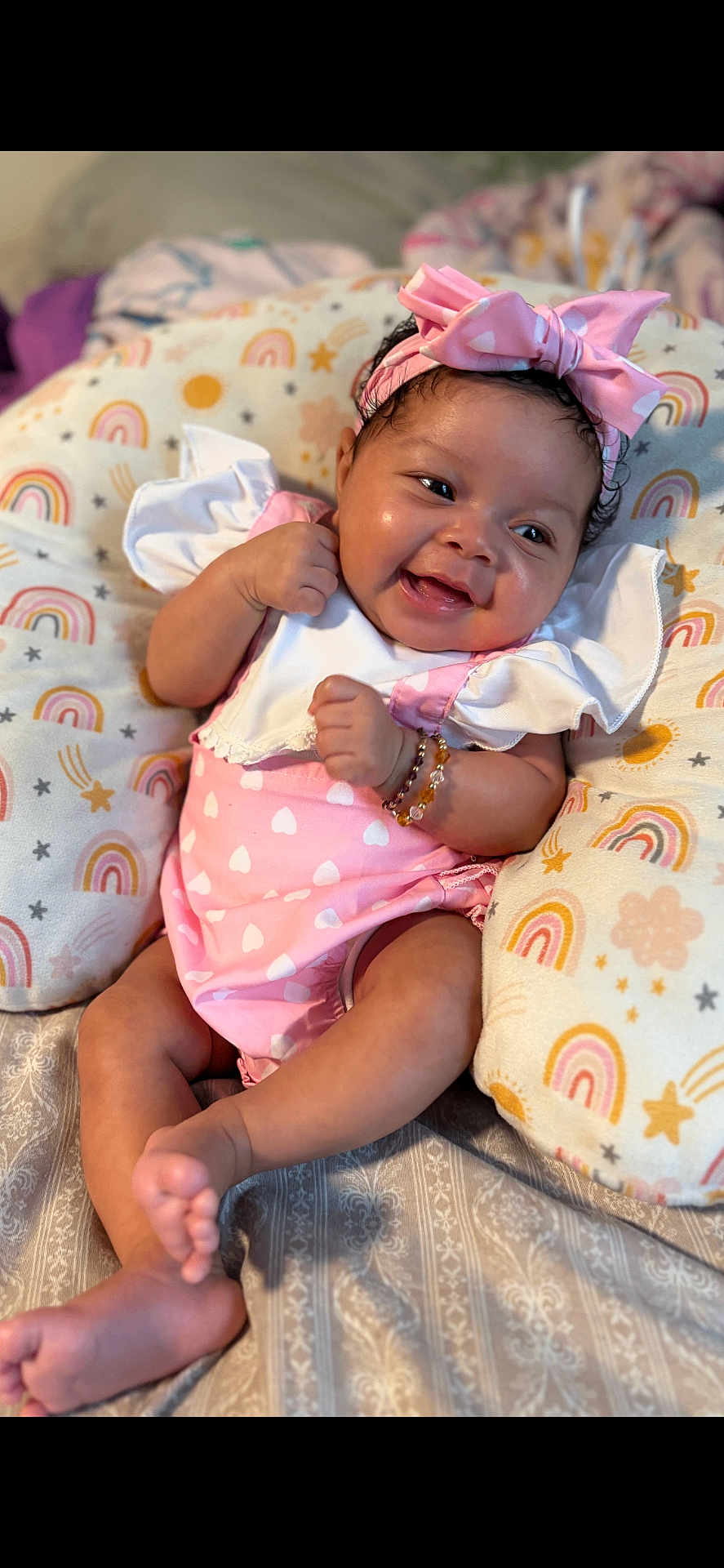 Azuri Howard is registered to the contest to win money with this photo: baby, bow, bracelet, child, close_up, cushion, cute, happy, headband, indoors, infant, lying_down, person, pink_clothing, rainbow_pattern, skin, smiling, soft_lighting, stars, toddler