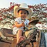 Liam is registered to the contest to win money with this photo: baby, child, cowboy_hat, saddle, rope, outdoor, tree, red_flowers, smiling, happy, person, sunset, nature, animal_saddle, foot, white_clothing, portrait, cute, young_child, daytime