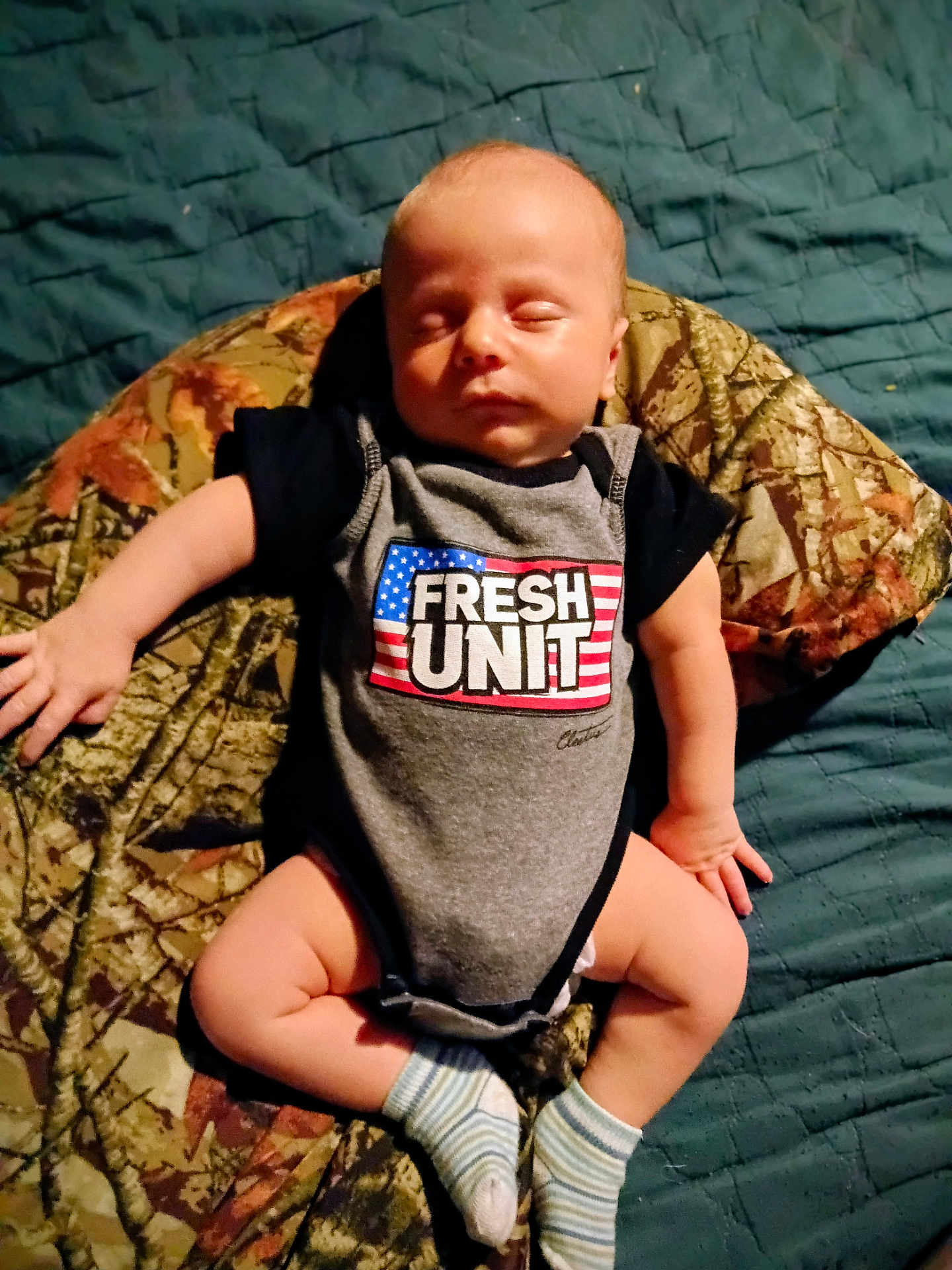 Dean is registered to the contest to win money with this photo: baby, sleeping, onesie, clothing, blanket, camouflage, blue, bed, infant, child, cute, peaceful, sock, lying_down, indoor, portrait, skin, newborn, hand, footwear