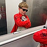 child, boy, sunglasses, red_jacket, mirror, reflection, elevator, pose, confident, casual_clothing, short_hair, indoor, young, standing, modern, smile, fashion, cool, hands_crossed, playful
