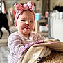 baby, child, pink_headband, bow, blanket, indoor, cozy, tongue_out, cute, portrait, soft_lighting, striped_clothing, infant, smiling, face, person, home, warm, closeup, sitting