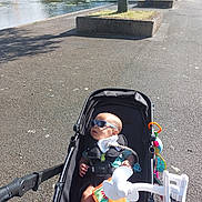 Wyatt is registered to the contest to win money with this photo: baby, stroller, sunglasses, outdoor, sunny, water, pavement, tree, toy, fan, child, shade, park, relaxation, infant, plaything, baby_bottle_holder, summer, leisure, person