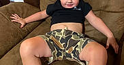 Aster is registered to the contest to win money with this photo: black_shirt, blonde, brown_furniture, camo_shorts, casual_clothing, chair, child, curly_hair, cute, face, hand, home, indoor, legs, relaxed, seated, skin, smile, sneakers, toddler