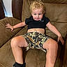 Aster is registered to the contest to win money with this photo: child, toddler, curly_hair, blonde, black_shirt, camo_shorts, sneakers, chair, brown_furniture, indoor, smile, relaxed, casual_clothing, cute, skin, face, legs, hand, seated, home