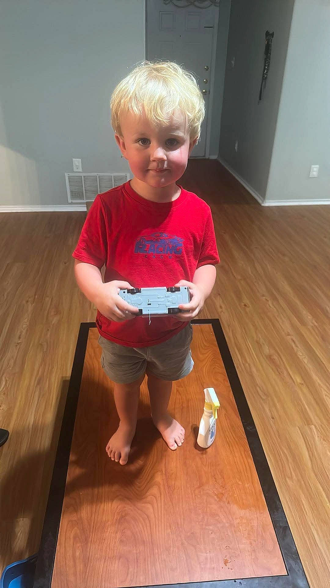 Aster is registered to the contest to win money with this photo: barefoot, blonde_hair, boy, casual_clothing, child, door, floor_mat, home, indoor, person, playtime, red_shirt, shorts, spray_bottle, standing, toy_car, vent, wall, wooden_floor, young_child