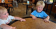 Aster is registered to the contest to win money with this photo: toddler, child, table, restaurant, indoor, blond_hair, blue_shirt, white_shirt, curious, playful, hands, chair, floor, window, natural_light, high_chair, brown_chair, tile_floor, smile, casual_clothing