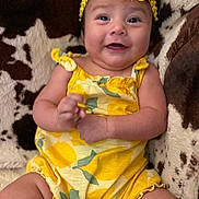 Leilani is registered to the contest to win money with this photo: baby, child, smiling, yellow_outfit, headband, flower, sitting, couch, patterned_fabric, happy, infant, cute, portrait, indoor, skin, arms, legs, comfort, clothing, person