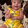 baby, child, smiling, yellow_outfit, headband, flower, sitting, couch, patterned_fabric, happy, infant, cute, portrait, indoor, skin, arms, legs, comfort, clothing, person