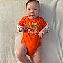 Lilith joined the competition — help win amazing prizes! baby, infant, onesie, orange_clothing, lying_down, blanket, cute, smiling, face, child, sweet, soft_texture, indoor, portrait, young_child, skin, hands, legs, pumpkin, expression