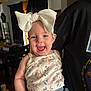 baby, child, smiling, bow, headwear, floral_top, denim_shorts, indoor, person, holding, happy, face, cute, infant, clothing, portrait, home, furniture, curtains, light