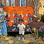 toddler, child, halloween, pumpkin, scarecrow, cornstalks, welcome_sign, flowers, hay, haunted_house, ghost, bat, brick_building, outdoor, costume, smiling, grass, daylight, festive, portrait