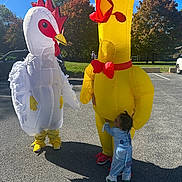 Carmelo joined the competition — help win amazing prizes! child, costume, inflatable, chicken, duck, parking_lot, outdoor, fall, trees, sunny, person, playful, hug, blue_onesie, yellow, white, red, green_shoes, shadow, fun