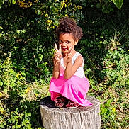 Malya participe au concours pour gagner de l'argent avec cette photo : child, girl, tree_stump, greenery, plants, sunlight, outdoor, nature, peace_sign, smiling, curly_hair, pink_skirt, sandals, flowers, grass, leaves, summer, portrait, playful, casual