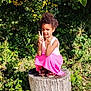 child, girl, tree_stump, greenery, plants, sunlight, outdoor, nature, peace_sign, smiling, curly_hair, pink_skirt, sandals, flowers, grass, leaves, summer, portrait, playful, casual