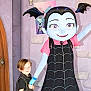 child, cartoon_character, vampire_costume, blue_skin, bat_wings, pigtails, dress, black_dress, pink_shirt, hand_holding, door, wall, poster, indoor, costume, young_child, short_hair, smile, friendly, character
