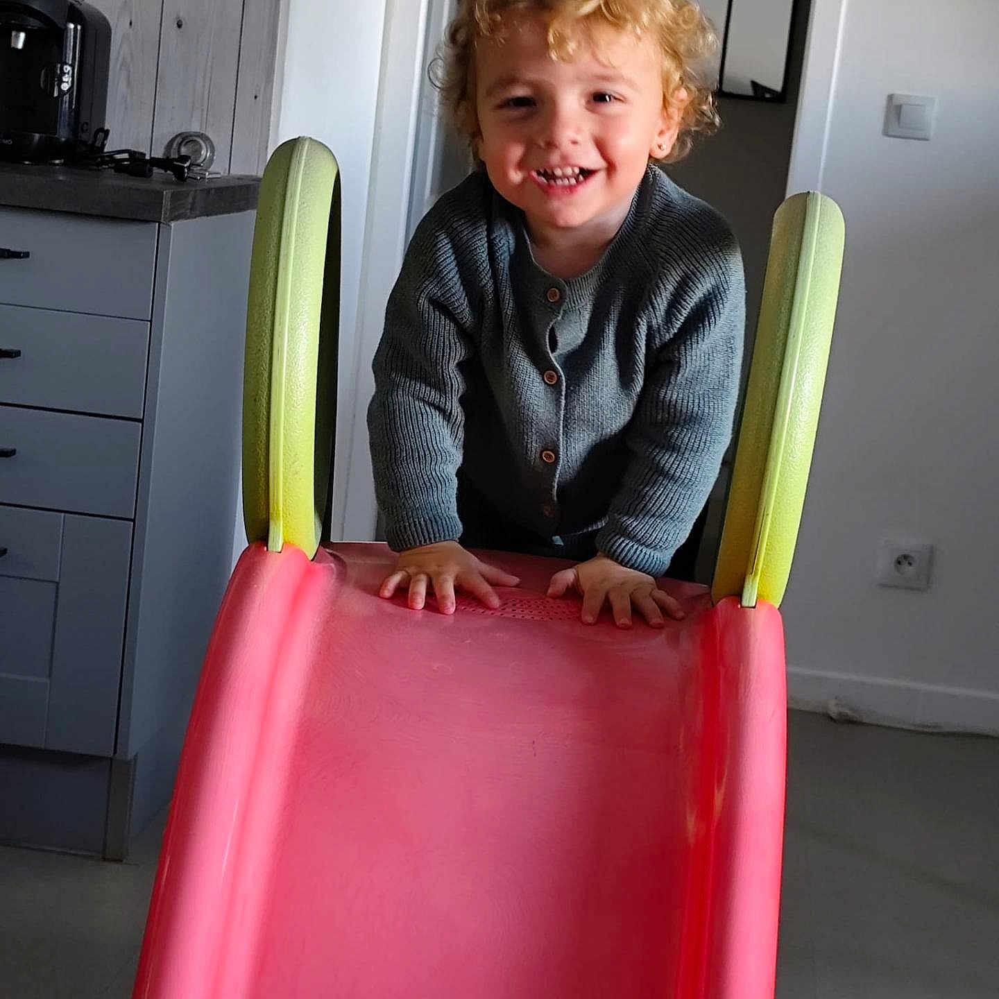 Kaylio Vinot participe au concours pour gagner de l'argent avec cette photo : cabinet, child, climbing, curly_hair, doorway, floor, grey_sweater, happy, home, indoor, kitchen, light_switch, person, pink_slide, play, slide, smiling, toddler, toy, young_child
