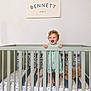 baby, banner, bed, blanket, child, crib, curly_hair, furniture, happy, home, interior, nursery, pattern, person, plush, room, smile, standing, toddler, toy