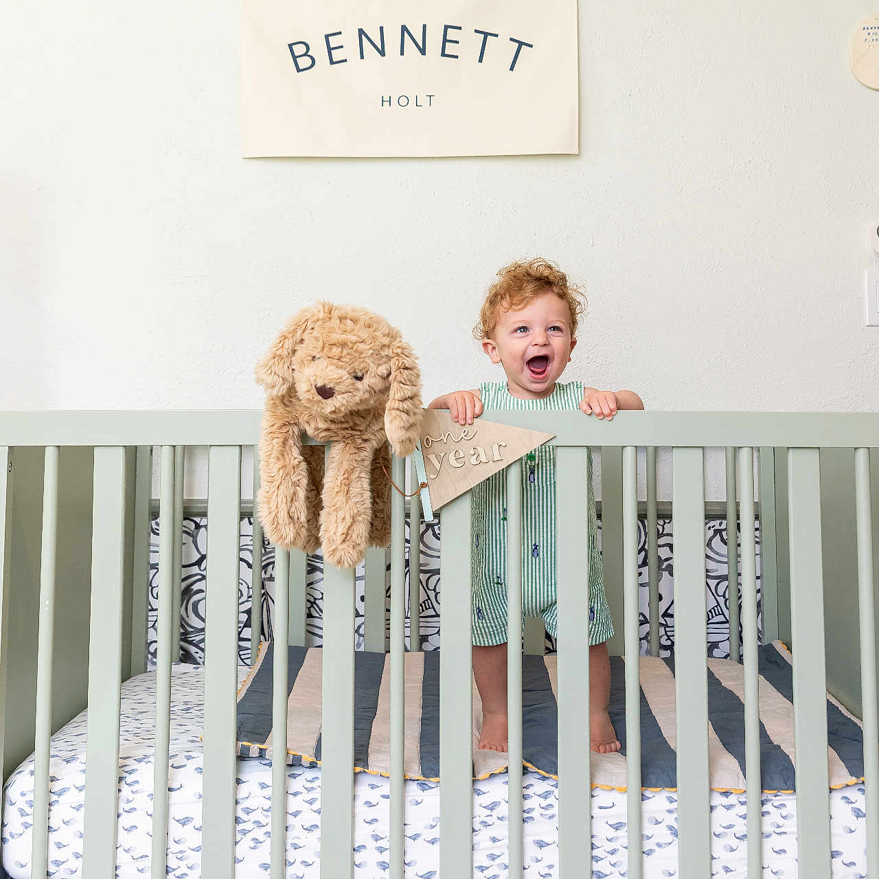 Bennett is registered to the contest to win money with this photo: baby, banner, bed, celebration, child, crib, curly_hair, decor, fabric, happy, home, indoor, nursery, one_year, person, smiling, standing, stuffed_animal, toddler, toy