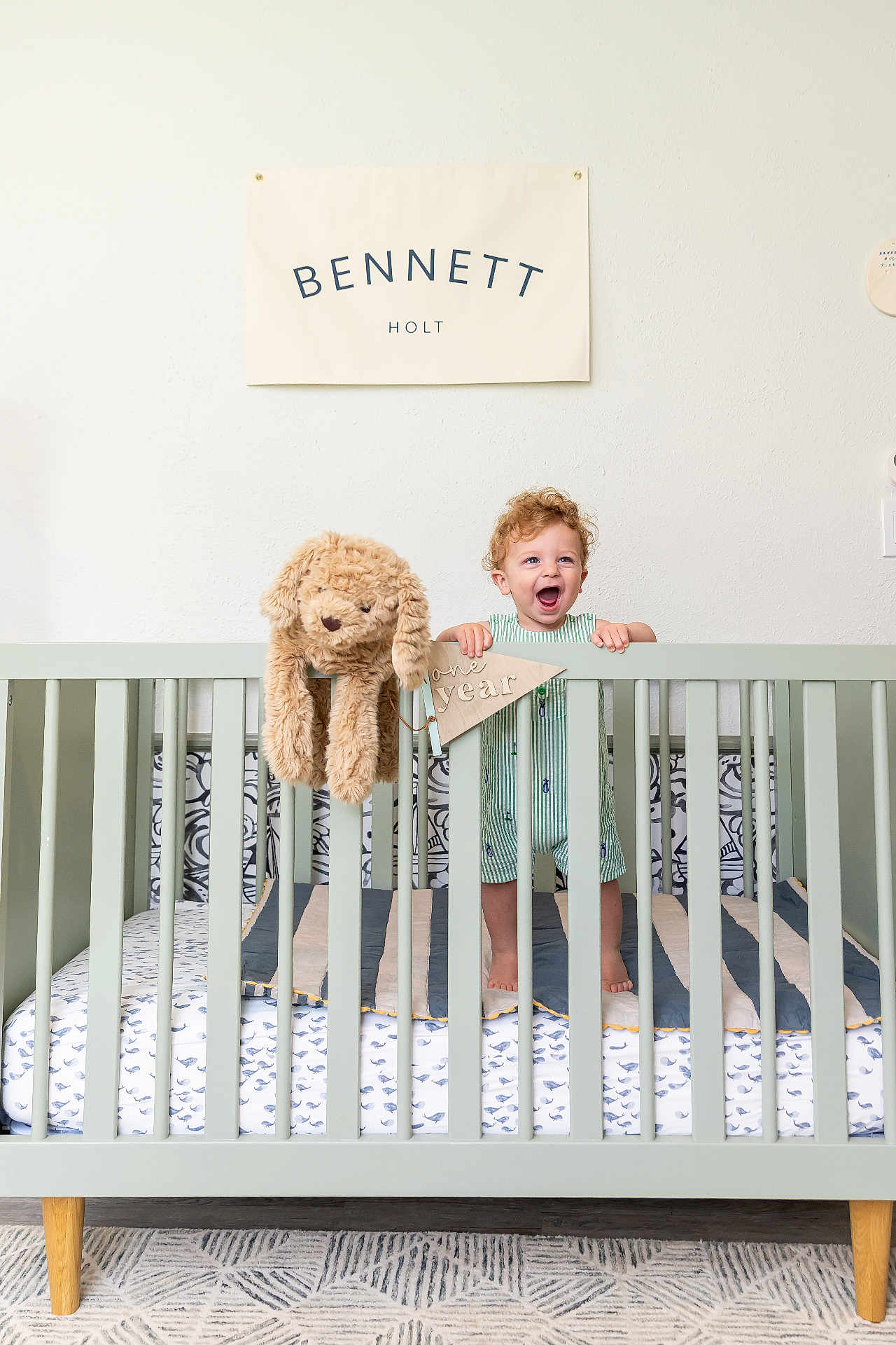 Bennett is registered to the contest to win money with this photo: baby, banner, bed, celebration, child, crib, curly_hair, decor, fabric, happy, home, indoor, nursery, one_year, person, smiling, standing, stuffed_animal, toddler, toy