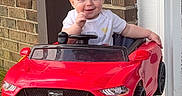 Dominic is registered to the contest to win money with this photo: baby, brick_wall, child, clothing, concrete_floor, cute, door, footwear_bare, front_view, happy, mustang, outdoor, person, red, smiling, steering_wheel, toy_car, vehicle_toy, walker, window