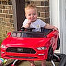 baby, toy_car, red, mustang, outdoor, brick_wall, door, happy, child, smiling, footwear_bare, steering_wheel, walker, vehicle_toy, person, concrete_floor, window, clothing, front_view, cute