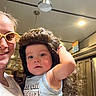 child, toddler, adult, person, wig, glasses, indoor, kitchen, smile, shirt, text, lighting, cabinet, ceiling_light, curly_hair, funny, portrait, holding, family, casual