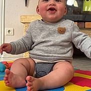 Thélyo participe au concours pour gagner de l'argent avec cette photo : baby, child, smiling, indoor, play_mat, sweater, teddy_bear_patch, toes, happy, floor, couch, fireplace, toy, cute, infant, person, head, face, legs, seated