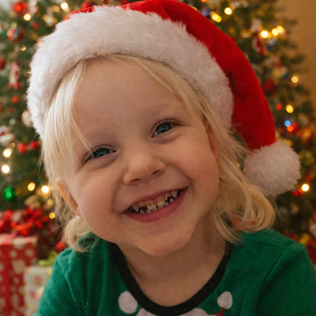 Selena is registered to the contest to win money with this photo: blonde_hair, celebration, child, christmas_lights, christmas_tree, cute, decorations, family, festive, green_sweater, happy, holiday, holiday_spirit, indoors, penguin, portrait, presents, santa_hat, seasonal, smiling