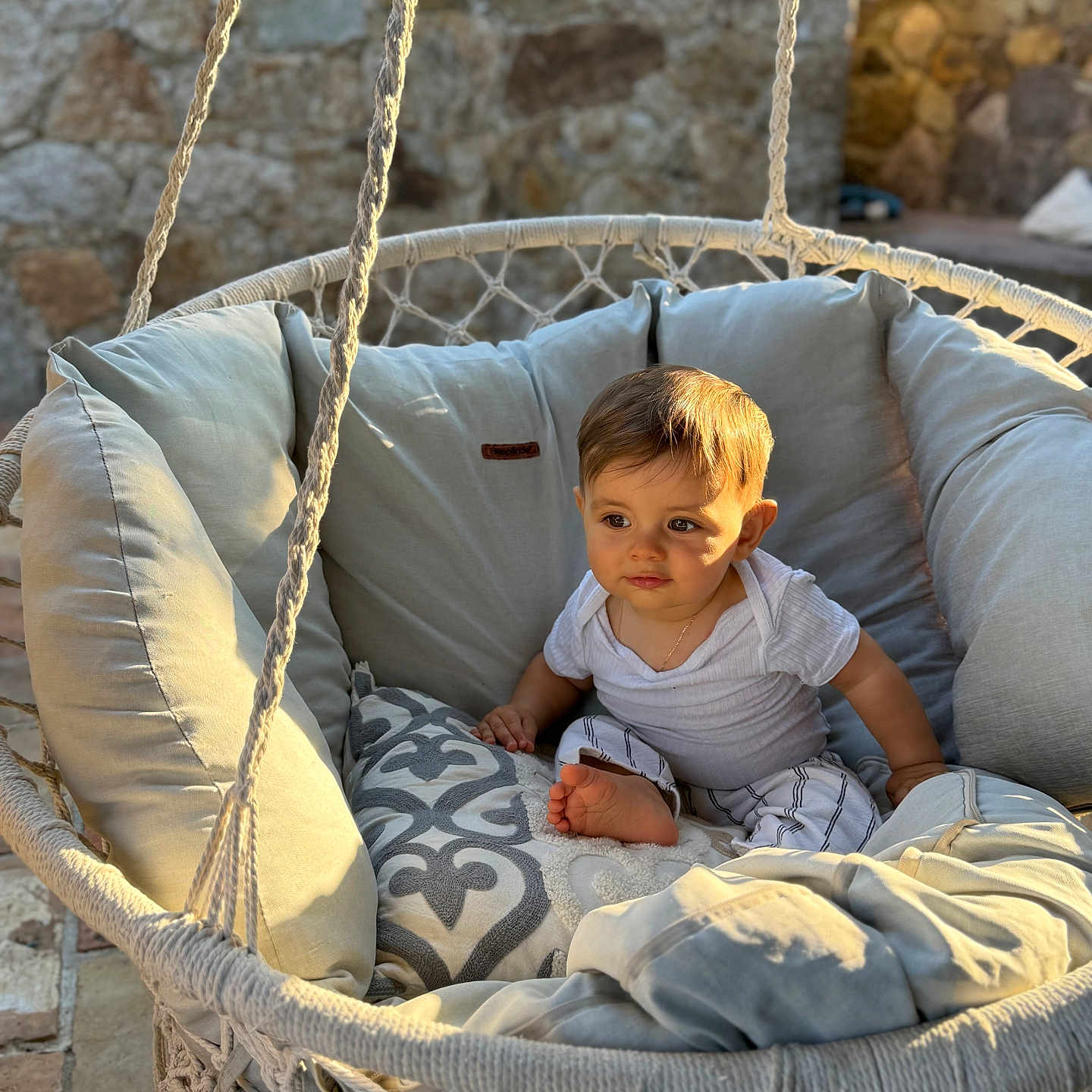 Ezio a rejoint le concours — aidez-le/la à gagner de superbes lots ! baby, child, cushion, cute, foot, hanging_chair, home_decor, infant, outdoor, pillow, portrait, relaxing, rope, seated, soft_furniture, stone_wall, striped_pants, sunlight, warm_light, white_clothing