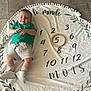 baby, child, green_shirt, beige_shorts, shoes, socks, round_blanket, numbers, wooden_ring, floor, milestone, age_marker, smiling, lying_down, indoor, decorative_blanket, text, french_words, young_child, person