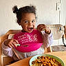 Eléna participe au concours pour gagner de l'argent avec cette photo : baby, bib, casual_clothing, child, curly_hair, cute, eating, food, happy, high_chair, indoor, meal, pasta, person, pigtails, pink_bib, plate, spoon, toddler, wooden_table