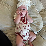 Blakely is registered to the contest to win money with this photo: baby, infant, child, headband, flower, onesie, cushion, carpet, hands_on_face, tongue_out, surprised, soft_fabric, cute, indoor, person, plush, skin, foot, leg, expression