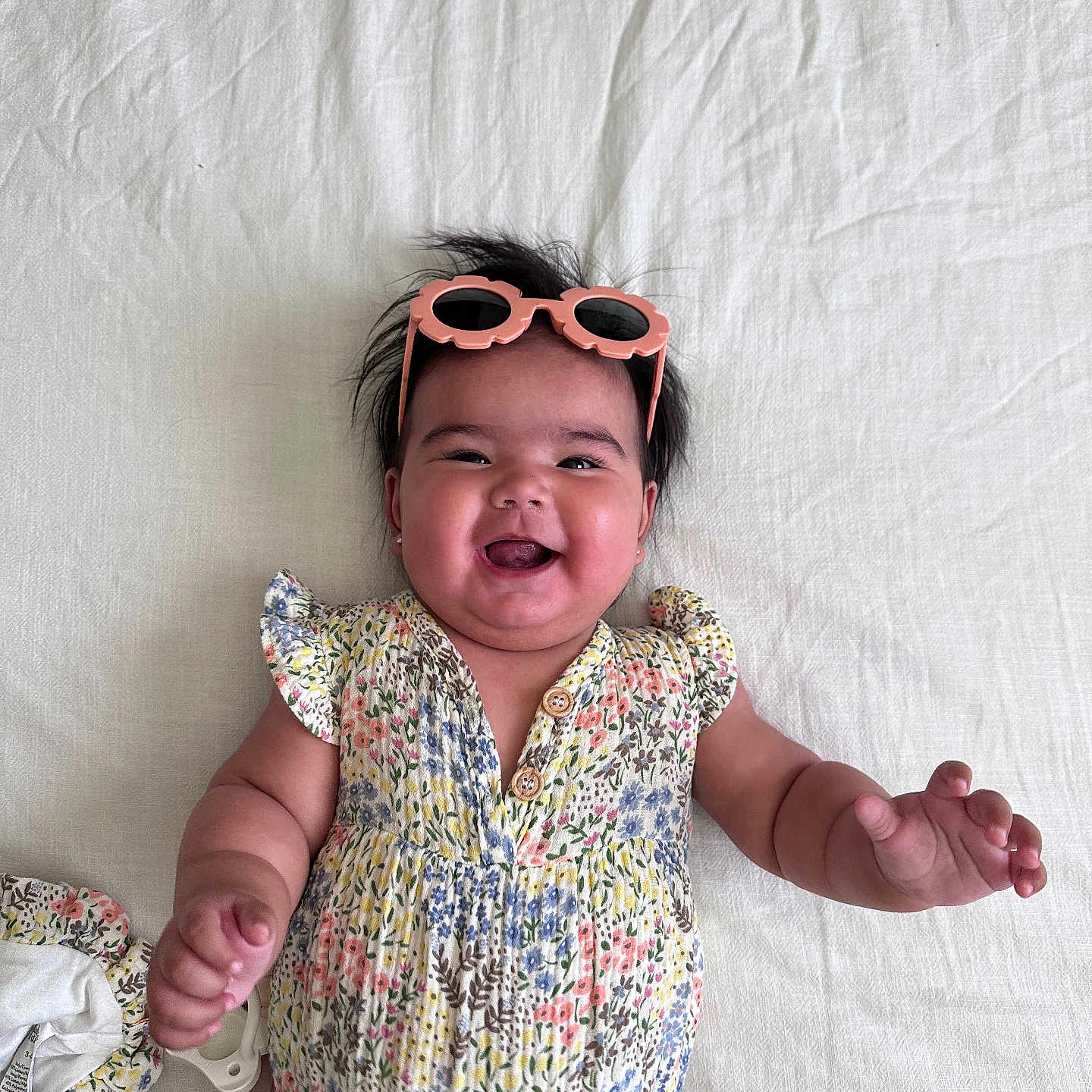 Saylah is registered to the contest to win money with this photo: accessory, baby, child, chubby, cute, earrings, fabric, face, floral_print, hand, happy, head, infant, lying_down, onesie, person, smiling, sunglasses, texture, white_background