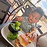 George joined the competition — help win amazing prizes! child, toddler, baby, curly_hair, smiling, face, high_chair, outdoor, restaurant, patio, food, burger, broccoli, meal, lunch, eating, plate, table, sunlight, portrait