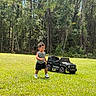 George joined the competition — help win amazing prizes! child, toddler, outdoor, grass, forest, trees, toy_car, toy_jeep, vehicle, walking, standing, striped_shirt, shorts, sneakers, sunlight, nature, lawn, portrait, playtime, adventure