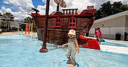 George joined the competition — help win amazing prizes! child, toddler, outdoor, splash_pad, water, playground, pirate_ship, swimsuit, sun_hat, striped_clothing, standing, vacation, pool, water_play, clouds, blue_sky, summer, architecture, adult, colorful