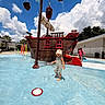 child, toddler, outdoor, splash_pad, water, playground, pirate_ship, swimsuit, sun_hat, striped_clothing, standing, vacation, pool, water_play, clouds, blue_sky, summer, architecture, adult, colorful