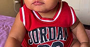 Kassio is registered to the contest to win money with this photo: baby, child, infant, red_clothing, jersey, jordan, number_23, sneakers, white_shoes, sitting, indoors, purple_blanket, curious_expression, cute, face, arm, legs, person, portrait, young