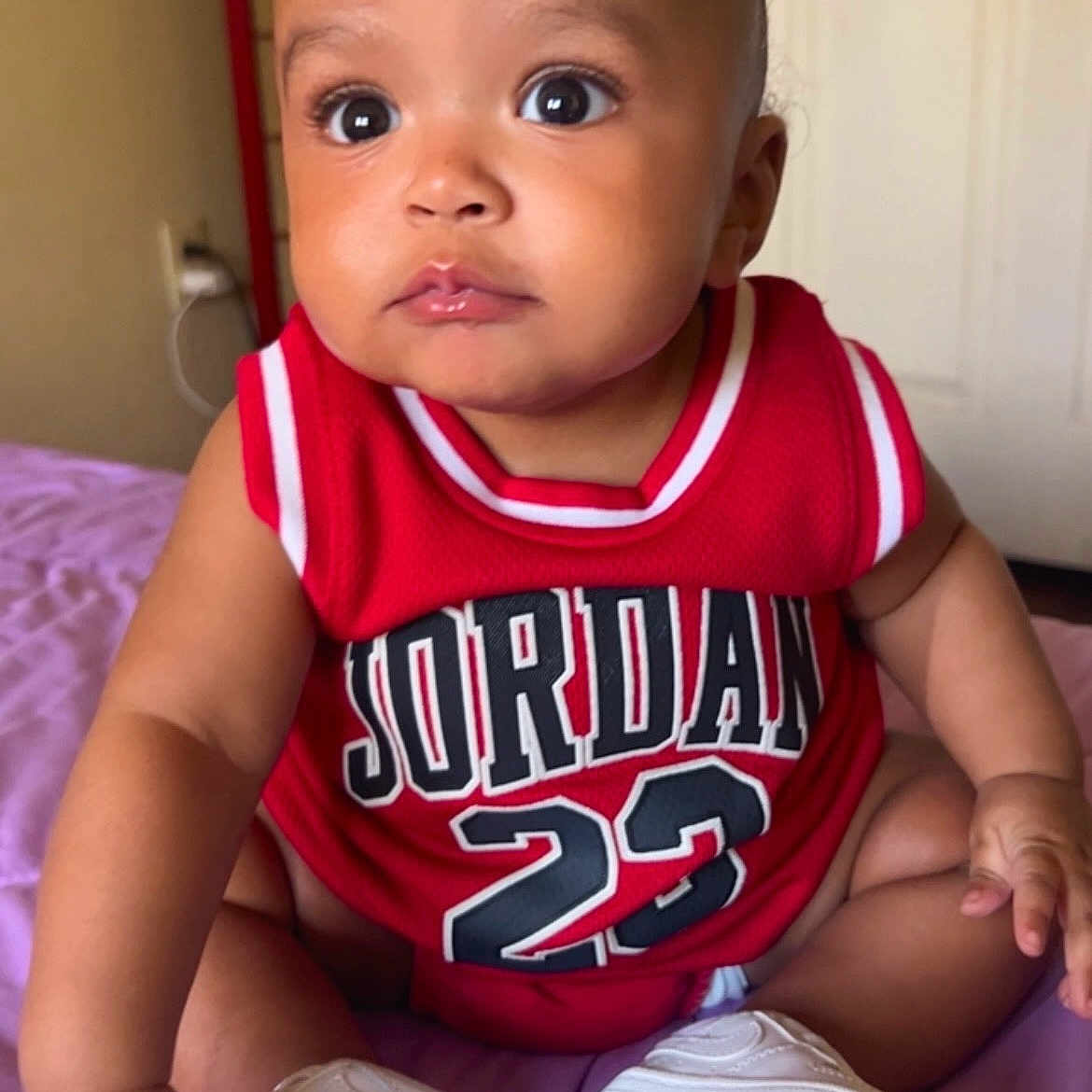 Kassio is registered to the contest to win money with this photo: baby, child, infant, red_clothing, jersey, jordan, number_23, sneakers, white_shoes, sitting, indoors, purple_blanket, curious_expression, cute, face, arm, legs, person, portrait, young