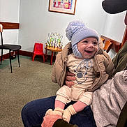 Riley is registered to the contest to win money with this photo: baby, child, smiling, hat, winter_clothing, indoor, adult, holding, seated, toy, chair, table, carpet, jacket, socks, face, person, happy, room, bead_maze
