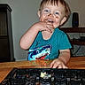 toddler, child, smiling, face, portrait, indoor, table, keyboard, cake, frosting, chocolate, messy, food, shark_shirt, turquoise_clothing, hand, sitting, home, photograph, playful
