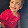 child, toddler, smiling, happy, portrait, seated, chair, indoor, pink_clothing, red_clothing, brown_hair, blue_eyes, face, teeth, arms, hands, casual, close_up, cozy, photography
